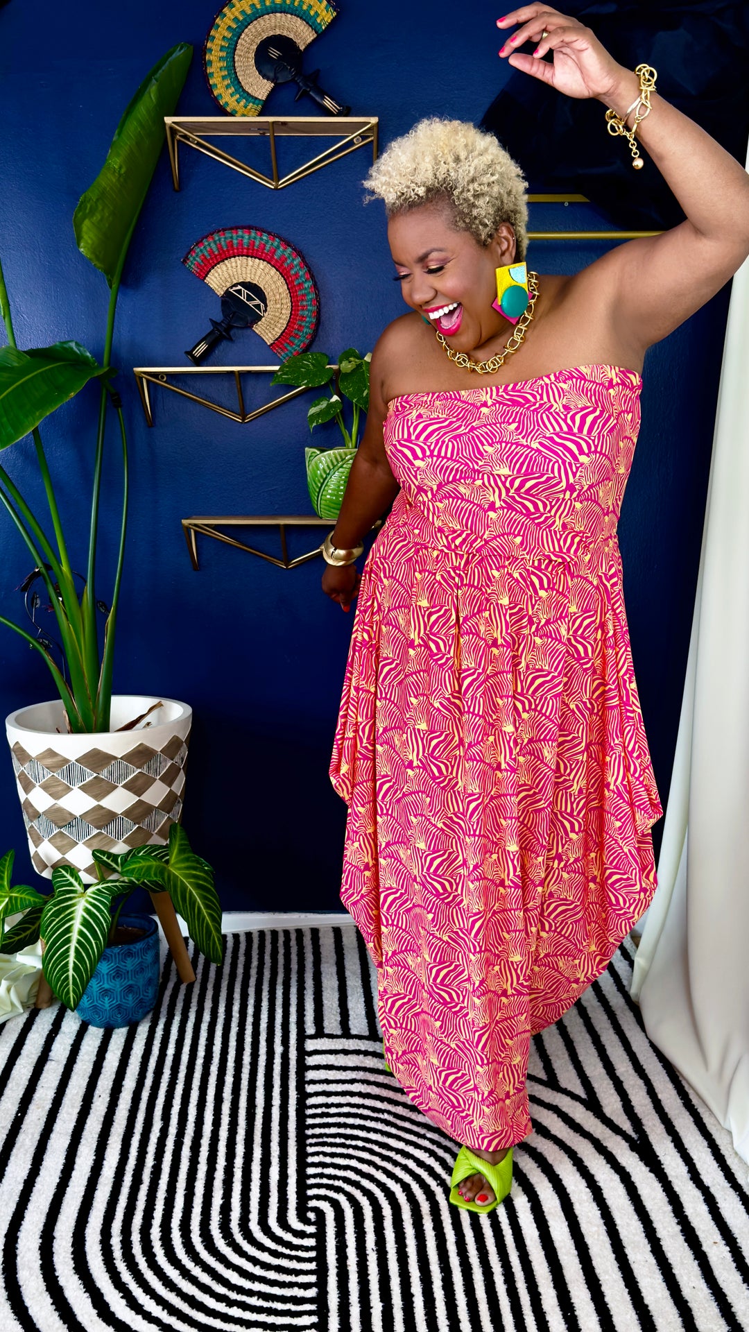 A woman modeling a strapless pink dress with a gold abstract pattern, standing in a styled room with a blue wall, gold shelves, plants, and a black-and-white patterned rug.