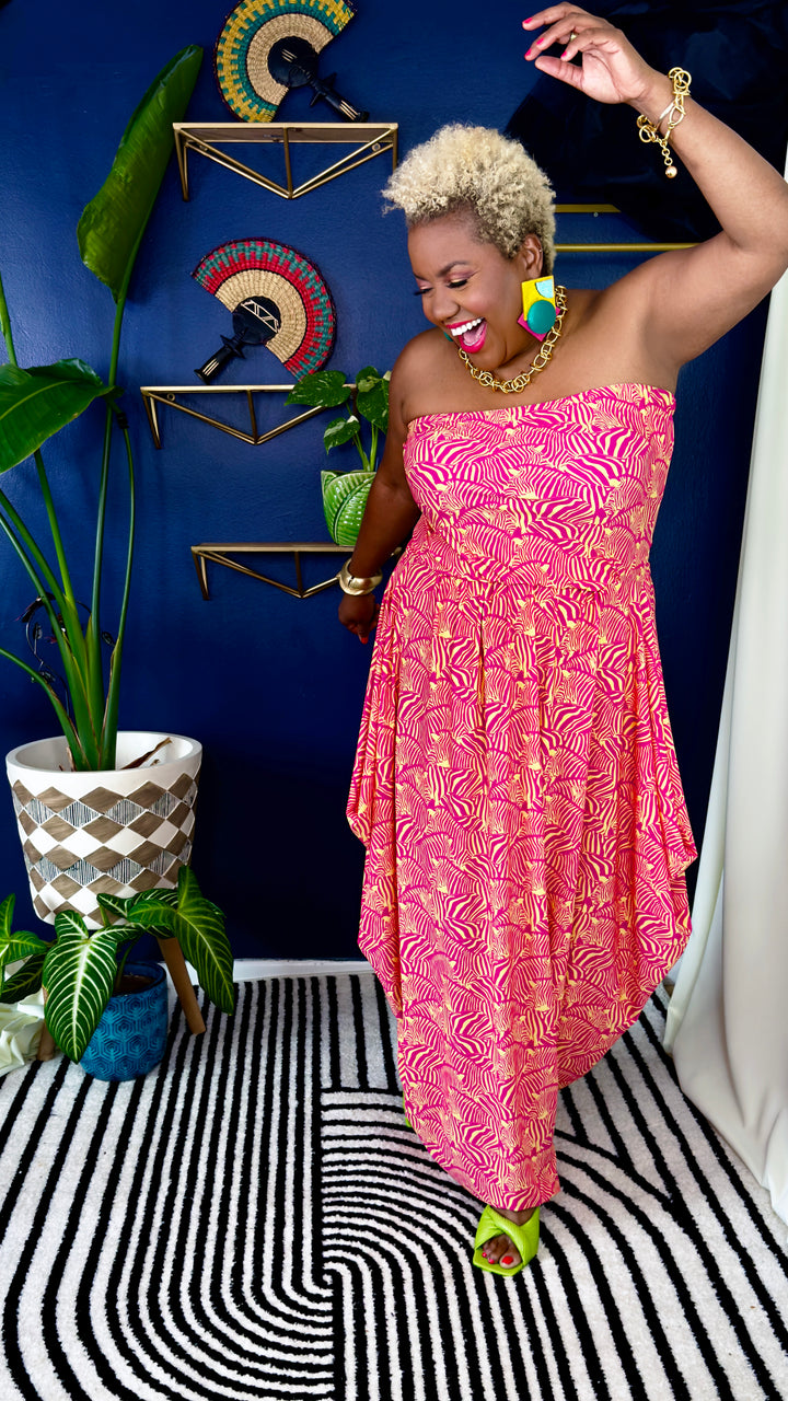 A woman modeling a strapless pink dress with a gold abstract pattern, standing in a styled room with a blue wall, gold shelves, plants, and a black-and-white patterned rug.