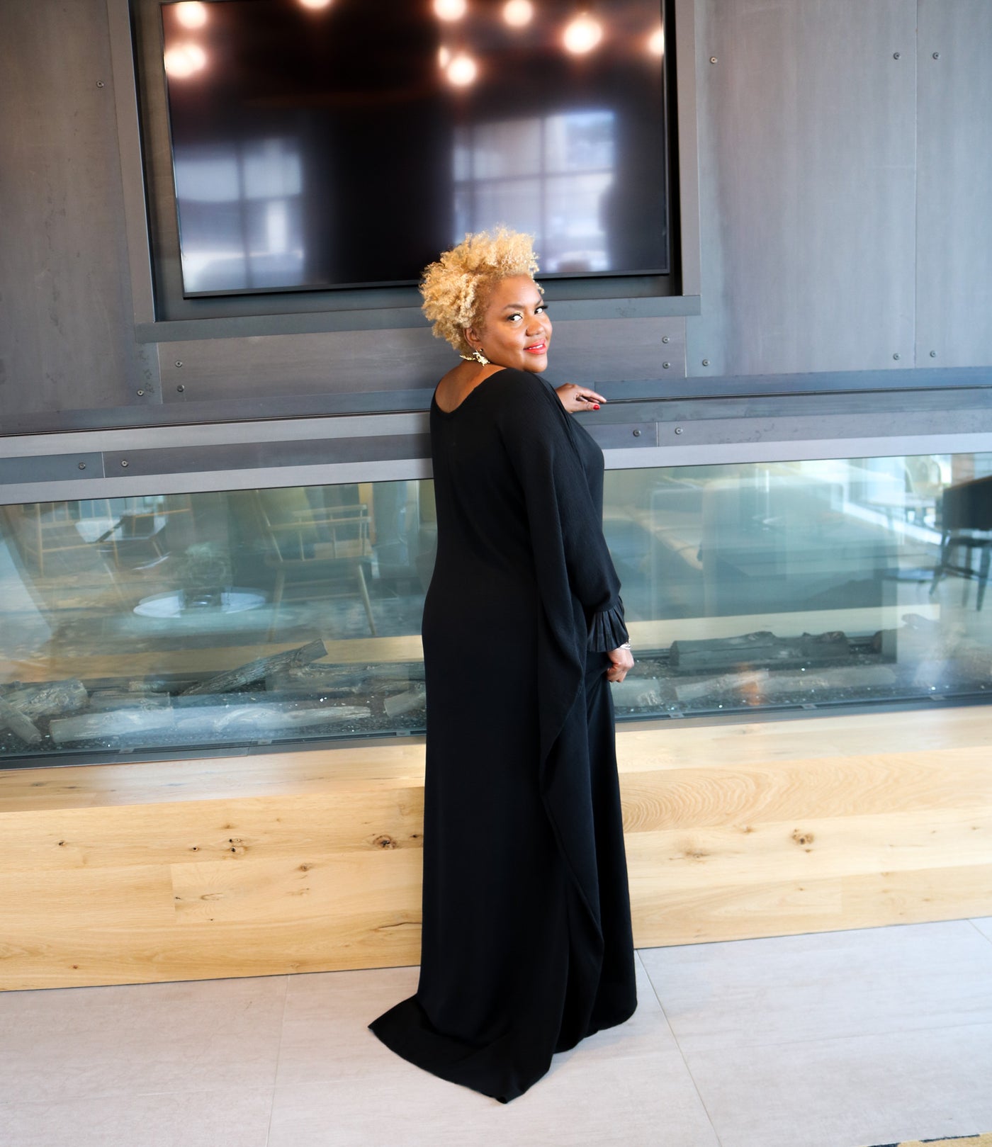 Woman in a black dress standing in a modern indoor setting with a large screen in the background.