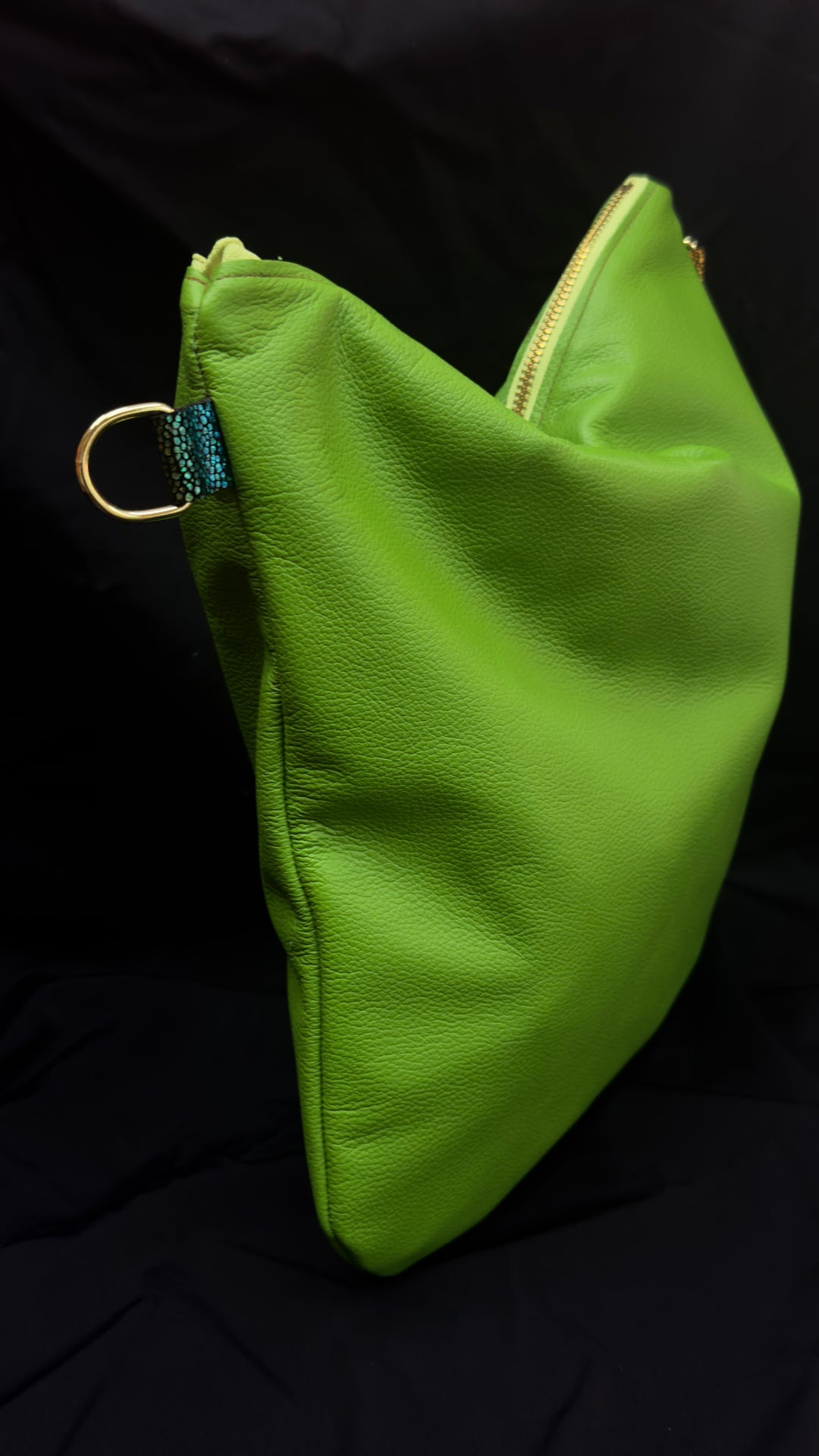 A bright lime green pebbled leather pouch bag with a gold zipper and a small gold-tone ring detail, shown against a black fabric background.
