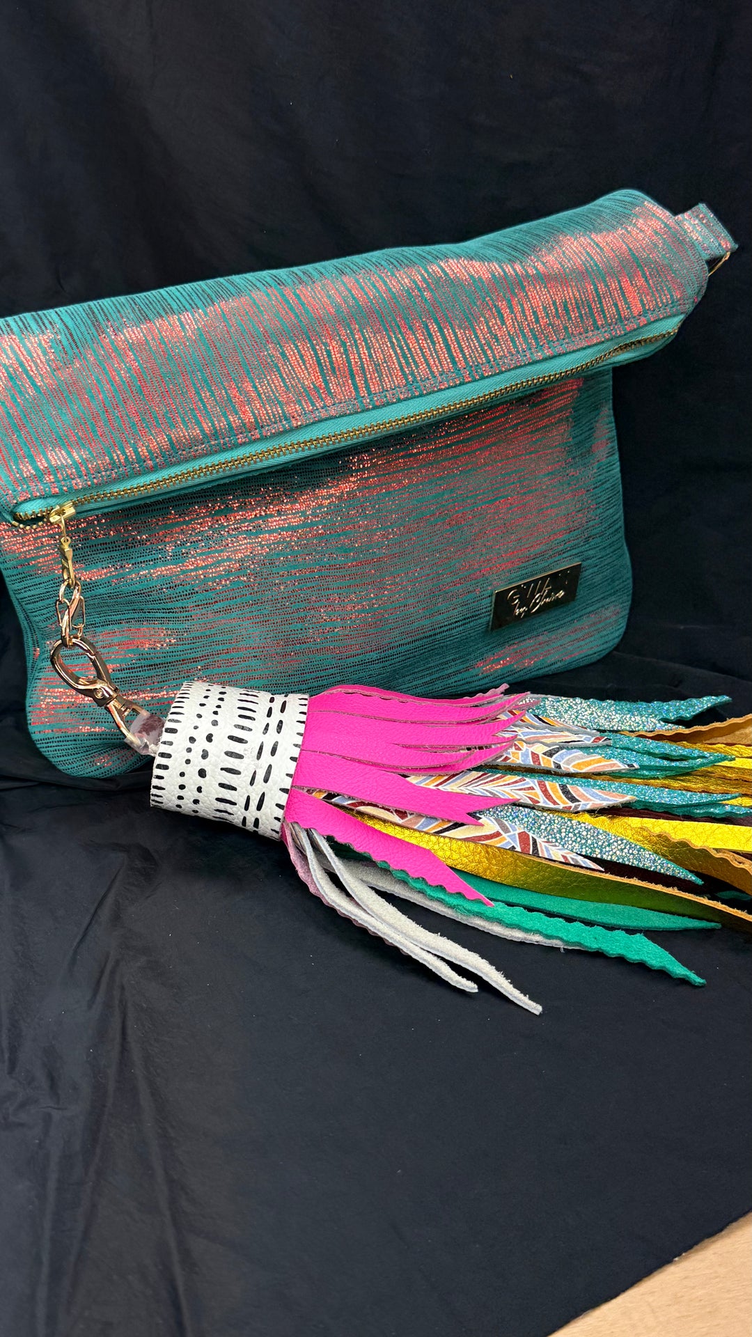 A teal and copper metallic clutch bag with a colorful tassel charm featuring pink, white, yellow, and green straps, displayed on a black fabric background.