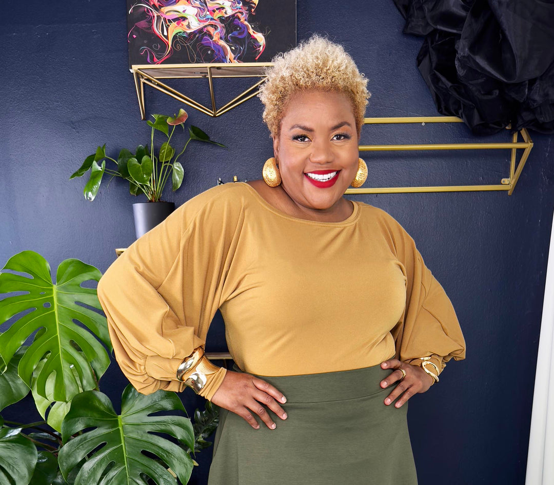Woman in mustard top and green skirt standing in a room with plants and decor.