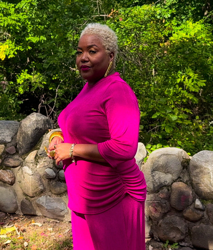 A woman with short gray hair wearing a vibrant magenta pink long-sleeve maxi dress stands outdoors in front of a rustic stone wall and lush green trees.
