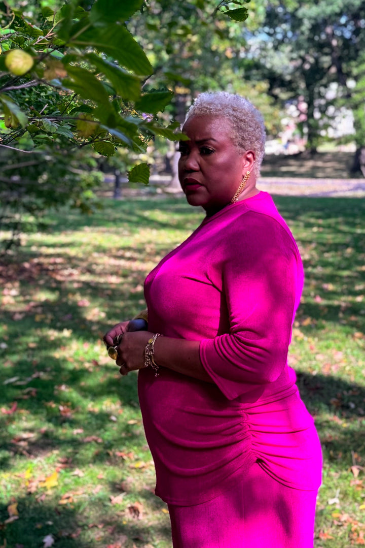A woman with short gray hair wearing a bright fuchsia pink long-sleeve ruched dress, standing outdoors in a grassy park area with trees and dappled sunlight.