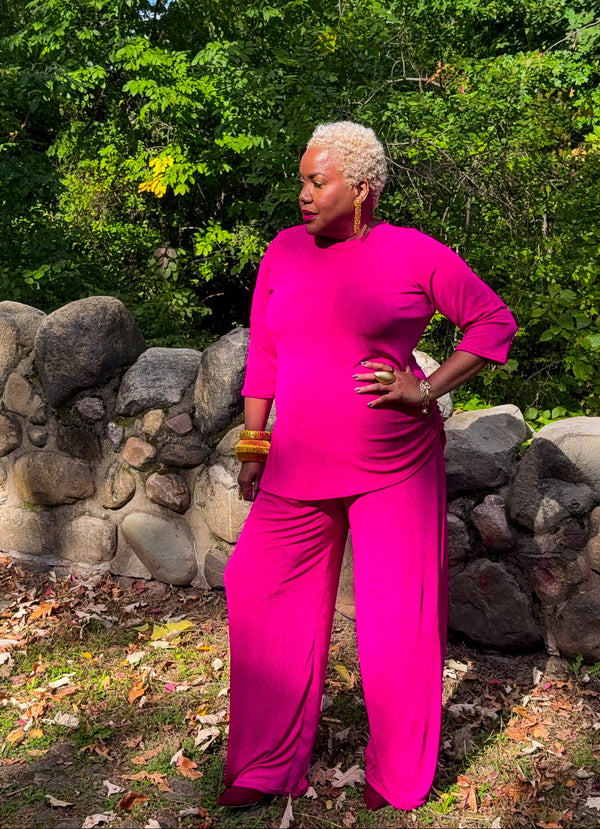 A woman with short blonde hair wearing a bright pink matching two-piece outfit stands outdoors in front of a stone wall and green trees. The outfit consists of a long-sleeve top and wide-leg pants.