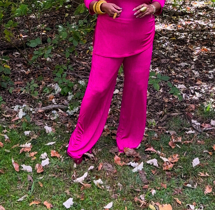 A woman with short blonde hair stands in a wooded outdoor setting wearing a bright pink matching two-piece outfit consisting of a long-sleeve top and wide-leg pants.
