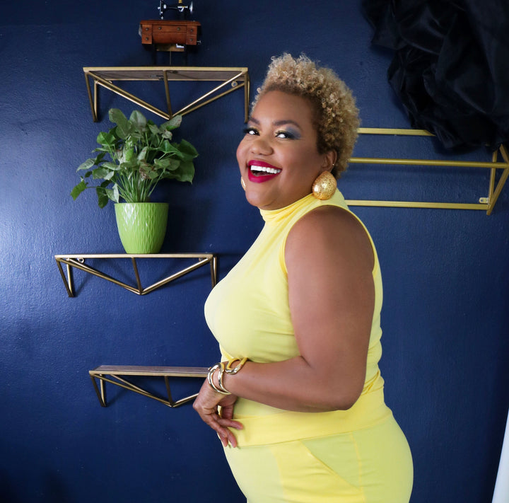 Woman in a yellow outfit standing in a room with a blue wall, black and white striped floor, and decorative shelves.
