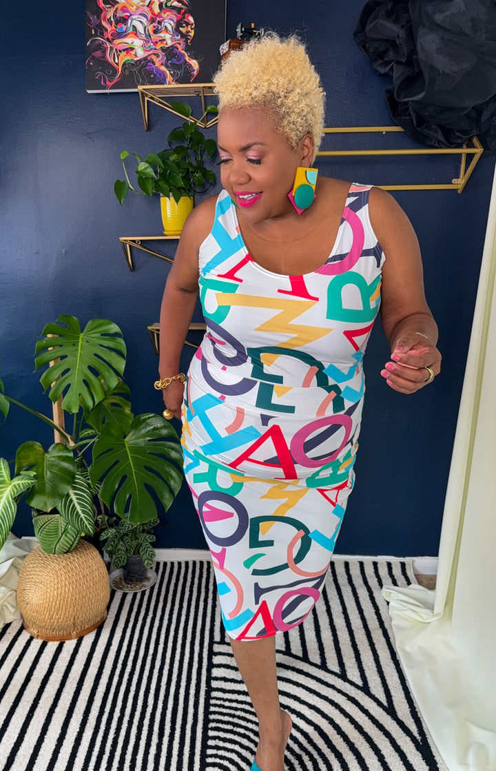Woman wearing a colorful dress with letter patterns in a room with plants and a blue wall.