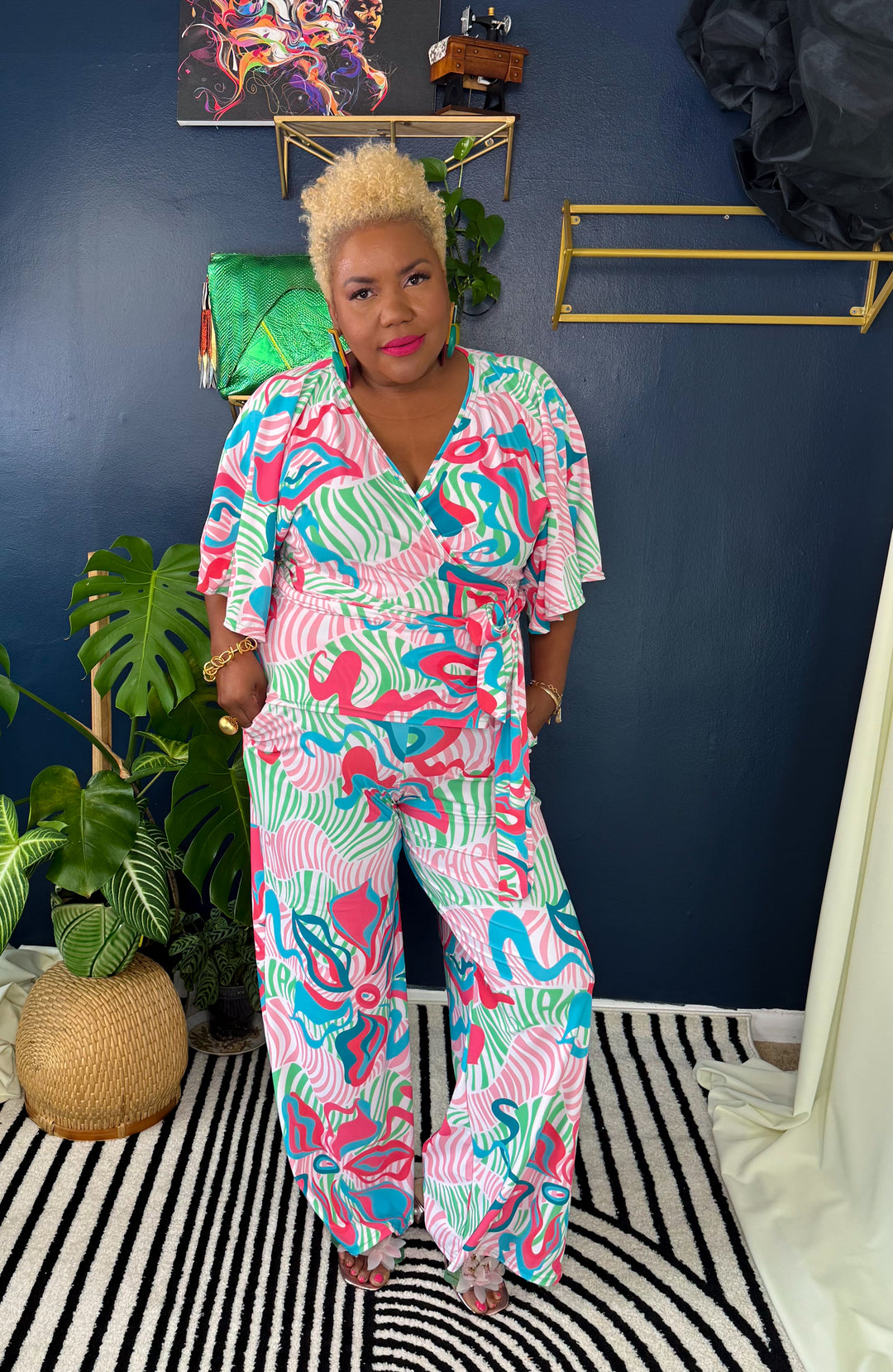 Woman in a colorful patterned outfit standing in a room with plants and decorative items.