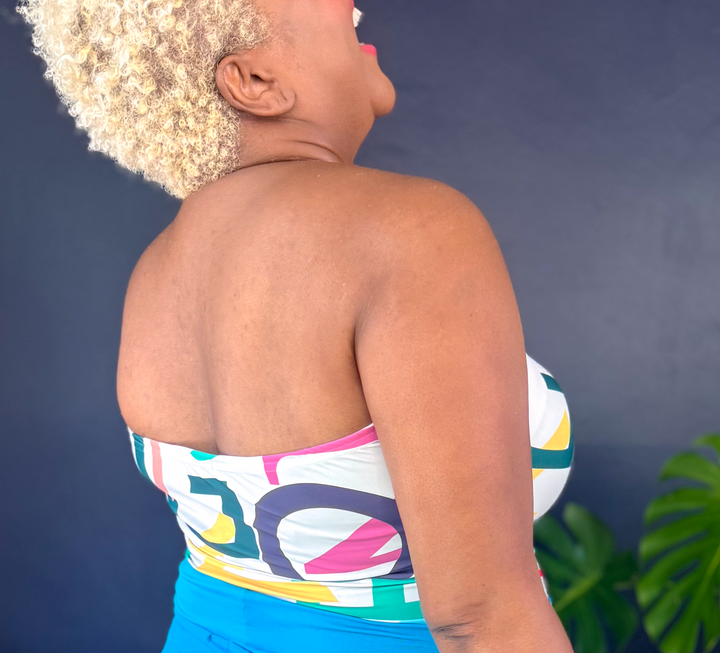A woman shown from the back wearing a strapless multicolor bandeau top and bright blue wide-leg pants, standing in a room with a dark blue wall, potted green plants, and a woven rug.