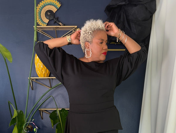 A woman with short platinum blonde hair models a black long-sleeve top and matching black pants in a styled indoor setting with a blue wall, plants, and a black-and-white patterned rug.