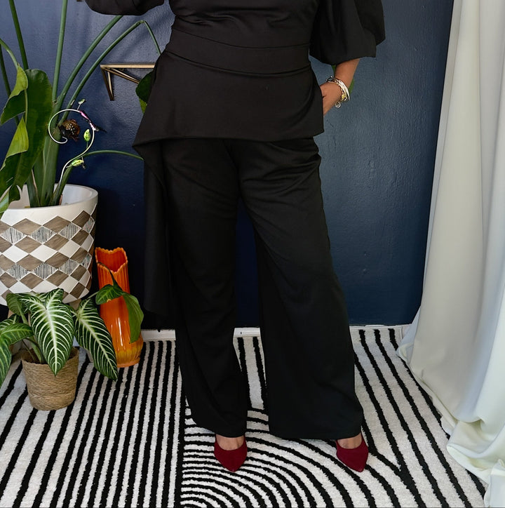 A woman modeling a black two-piece outfit consisting of a peplum-style top with voluminous puff sleeves and matching wide-leg trousers, standing in a styled room with a blue wall, plants, and a black-and-white striped rug.