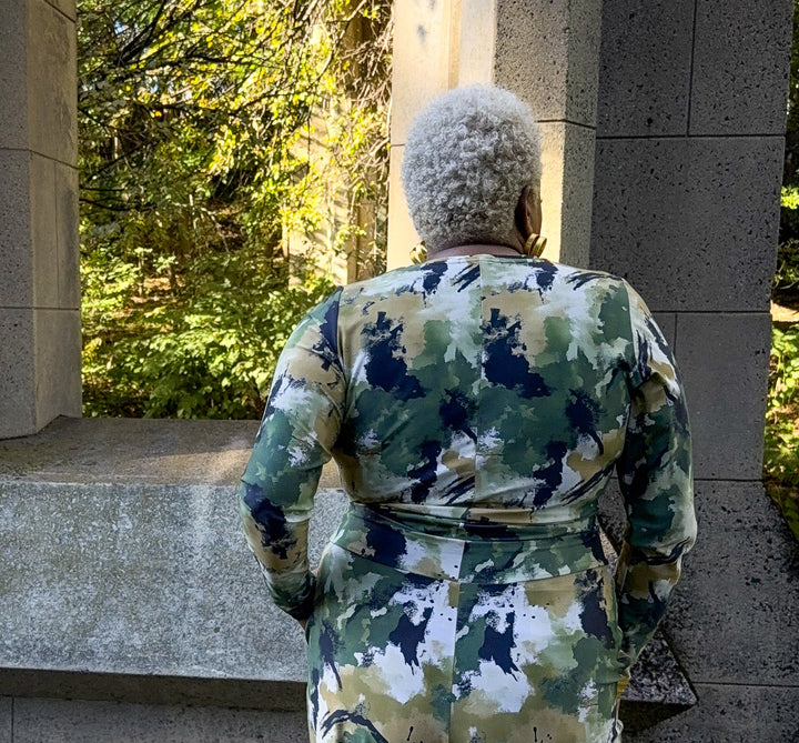 A person viewed from behind wearing a camouflage jumpsuit outdoors on a paved walkway near stone steps and greenery.