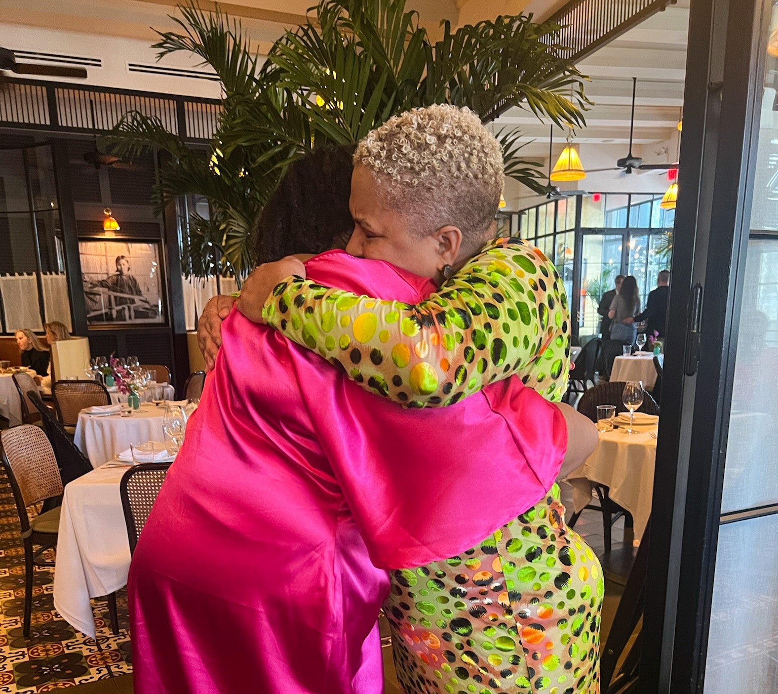 Two women hugging in a restaurant setting with tables and chairs visible.