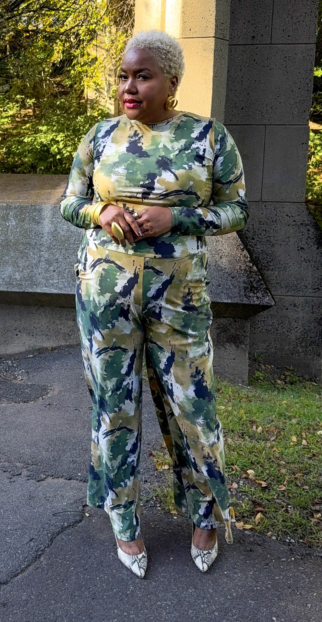 A woman wearing a matching long-sleeve top and wide-leg pants set in a green, beige, and navy camouflage pattern, standing outdoors on a paved path.