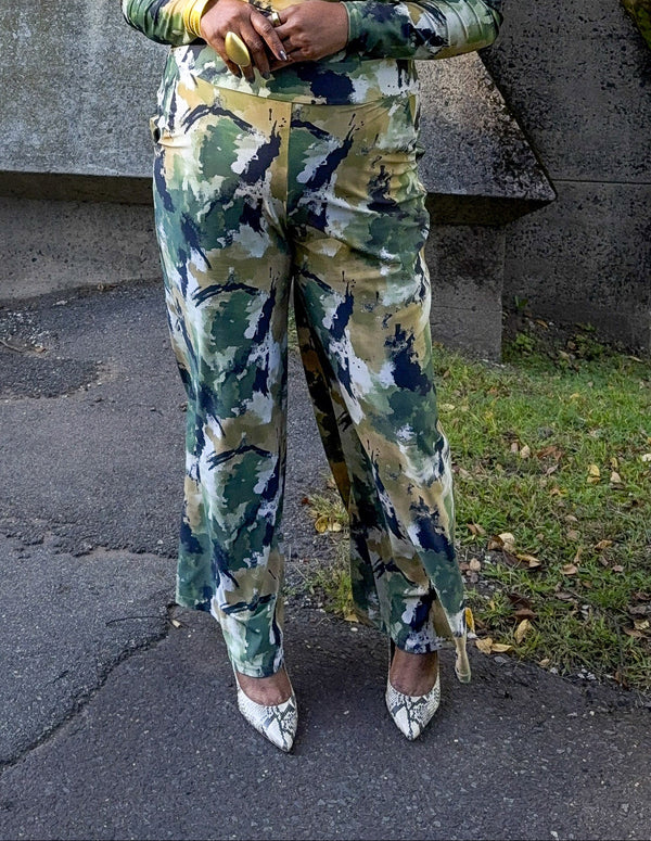 A woman wearing a matching long-sleeve top and wide-leg pants set in a green, beige, and navy camouflage pattern, standing outdoors on a paved path.