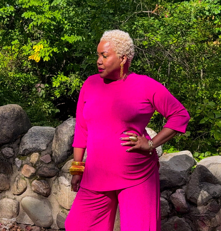 A woman with short blonde hair wearing a bright pink matching two-piece outfit stands outdoors in front of a stone wall and green trees. The outfit consists of a long-sleeve top and wide-leg pants.