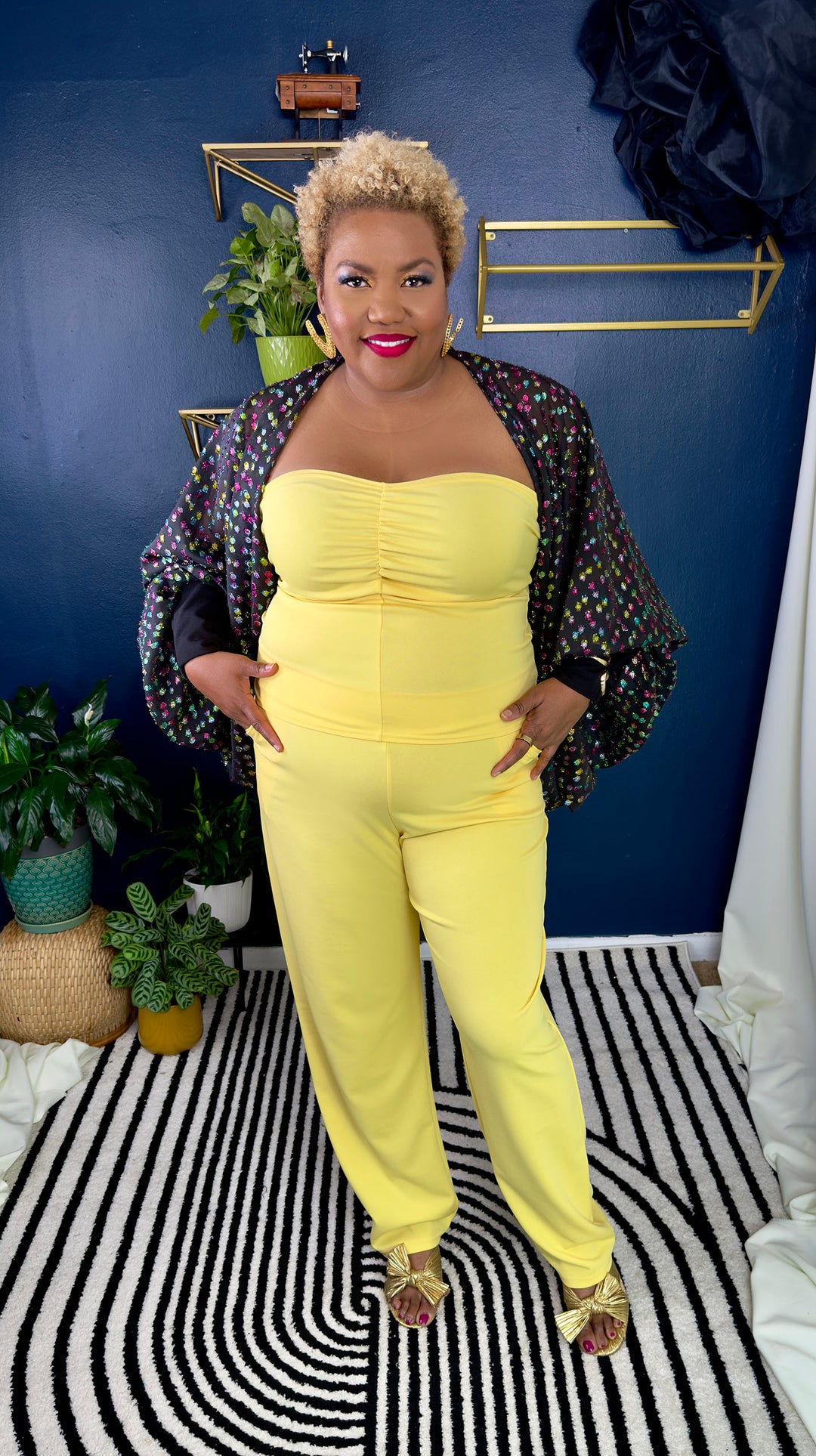 Woman in a yellow outfit standing on a black and white striped rug with a blue wall and plants in the background.