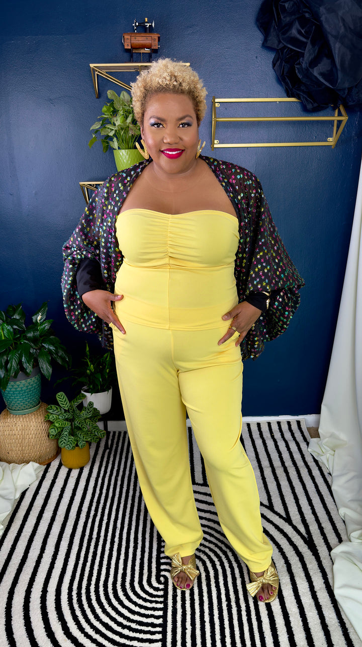 Woman in a yellow outfit standing on a black and white striped rug with a blue wall and plants in the background.