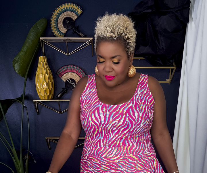 A woman wearing a sleeveless pink and white zebra-print peplum top and matching skirt, walking in a styled indoor setting with a black and white patterned rug, dark blue wall, and decorative shelves.